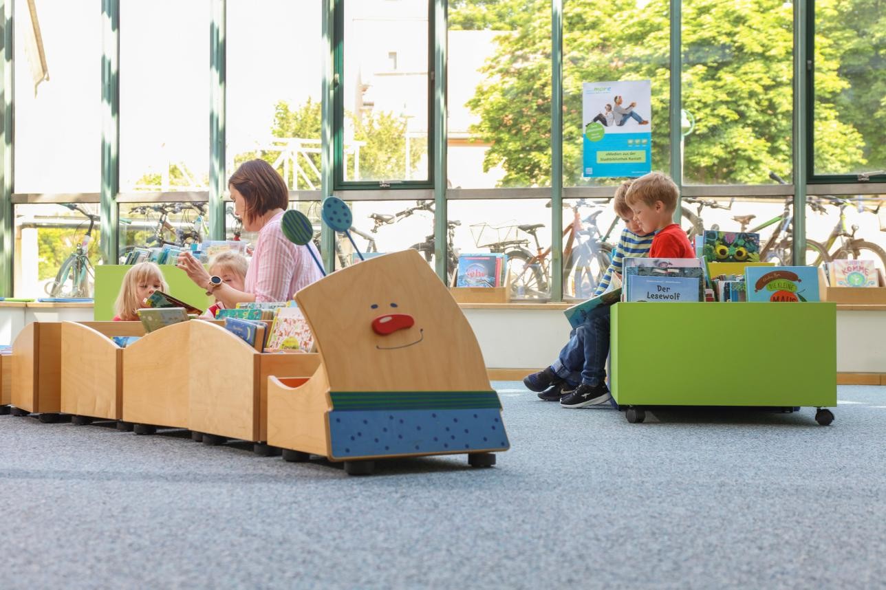 Children's area of the library with boxes of books and seating, children and a mother sit there and read.