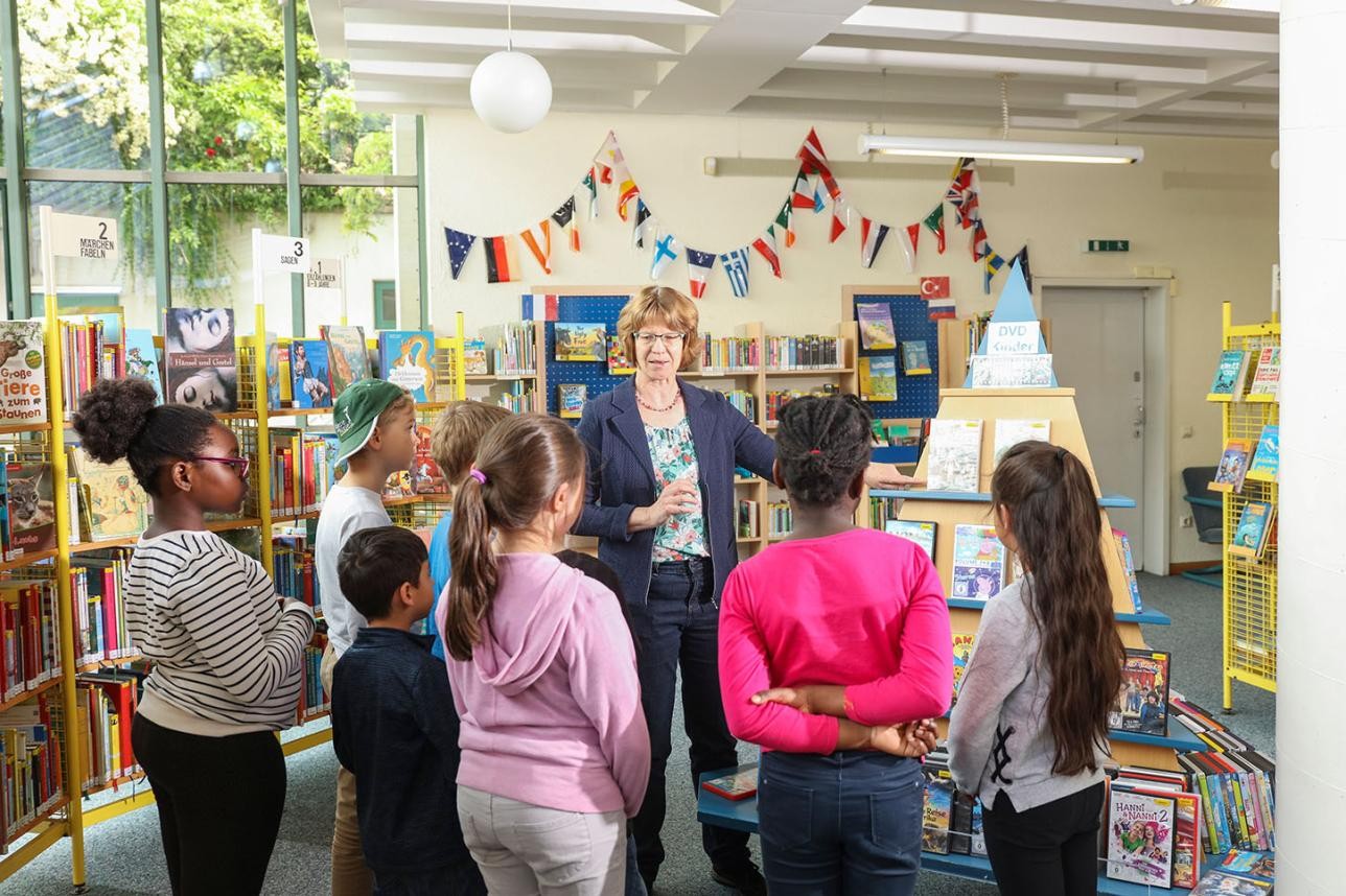 Bibliotheksführung - mehrere Kinder stehen um Birgitt Stellmach herum, während Sie etwas erzählt. Im Hintergrund Bücherregale.