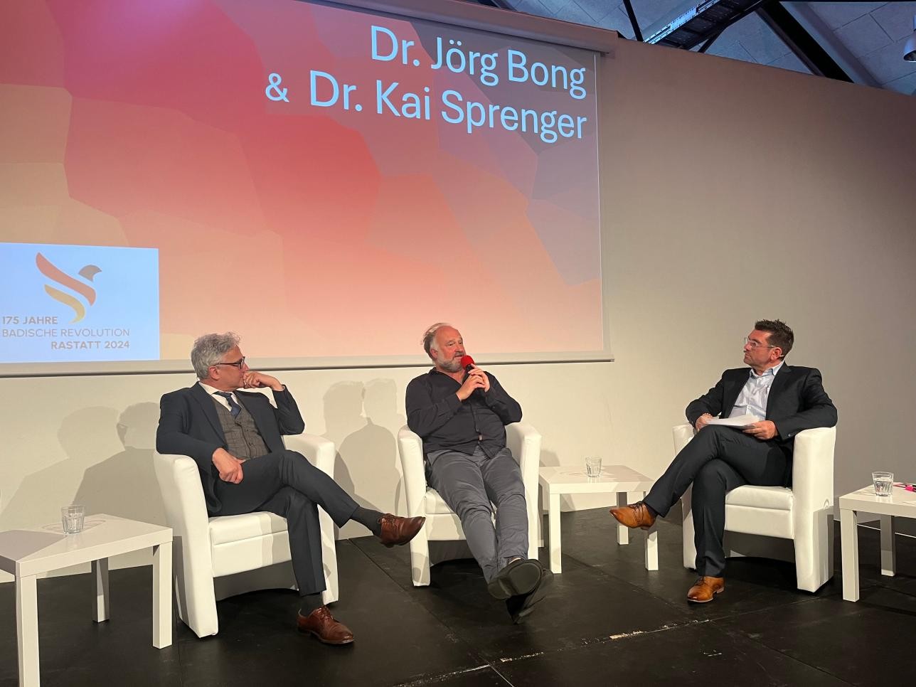 Dr. Kai Sprenger, Dr. Jörg Bong und Moderator Moderator Jochen Graf auf der Bühne in der Reithalle bei einer Diskussionsrunde