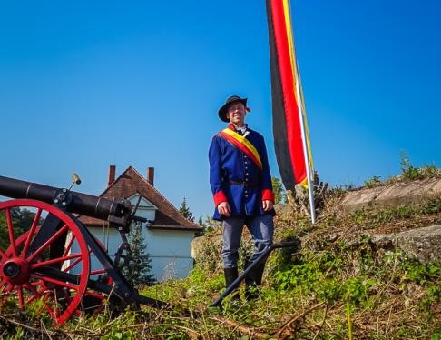 Schauspieler in historischem Kostüm steht vor einer Fahne und einer Kanone