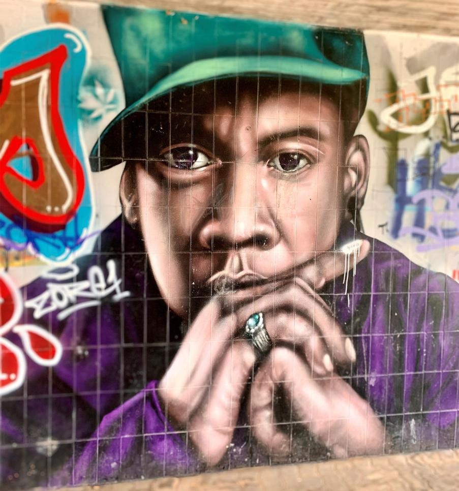 Wall sprayed with colorful graffiti, head, shoulders and hands of a man with a green cap, purple jacket and finger ring. His head is leaning on his folded hands.