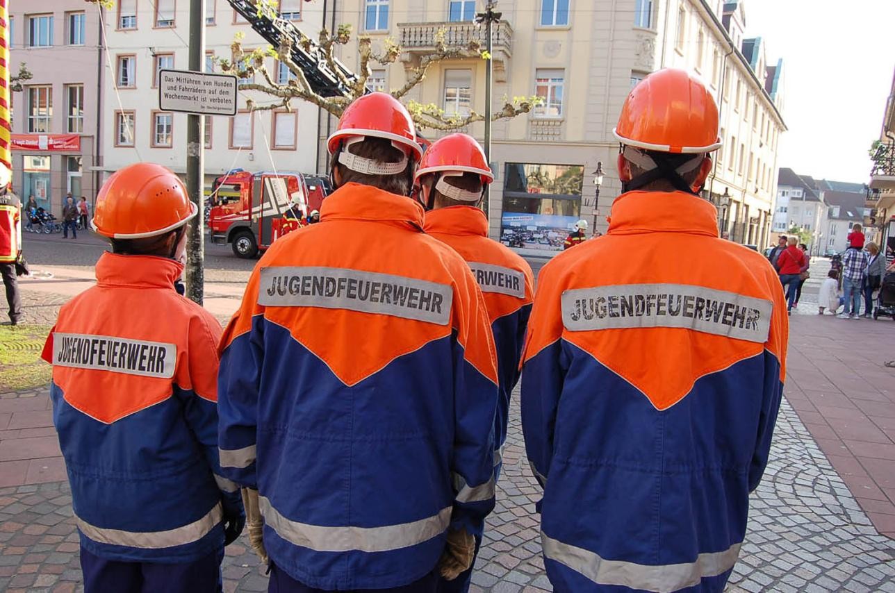 Four young people in firefighters uniform from behind, in the background houses, streets, spectators, a turntable ladder truck of the fire department