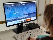 Bildschirm mit Tastatur, eine Frau von hinten, die mit einem Stift in der Hand am Schreibtisch sitzt. Der Bildschirm zeigt ein großes Bild von einem Gletscher.