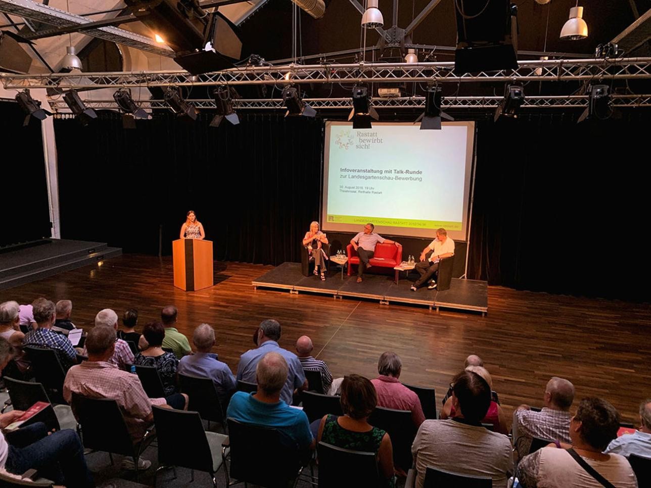 Informationsveranstaltung zur Landesgartenschau-Bewerbung. Redner sitzen auf der Bühne und Publikum befindet sich  in der Halle.