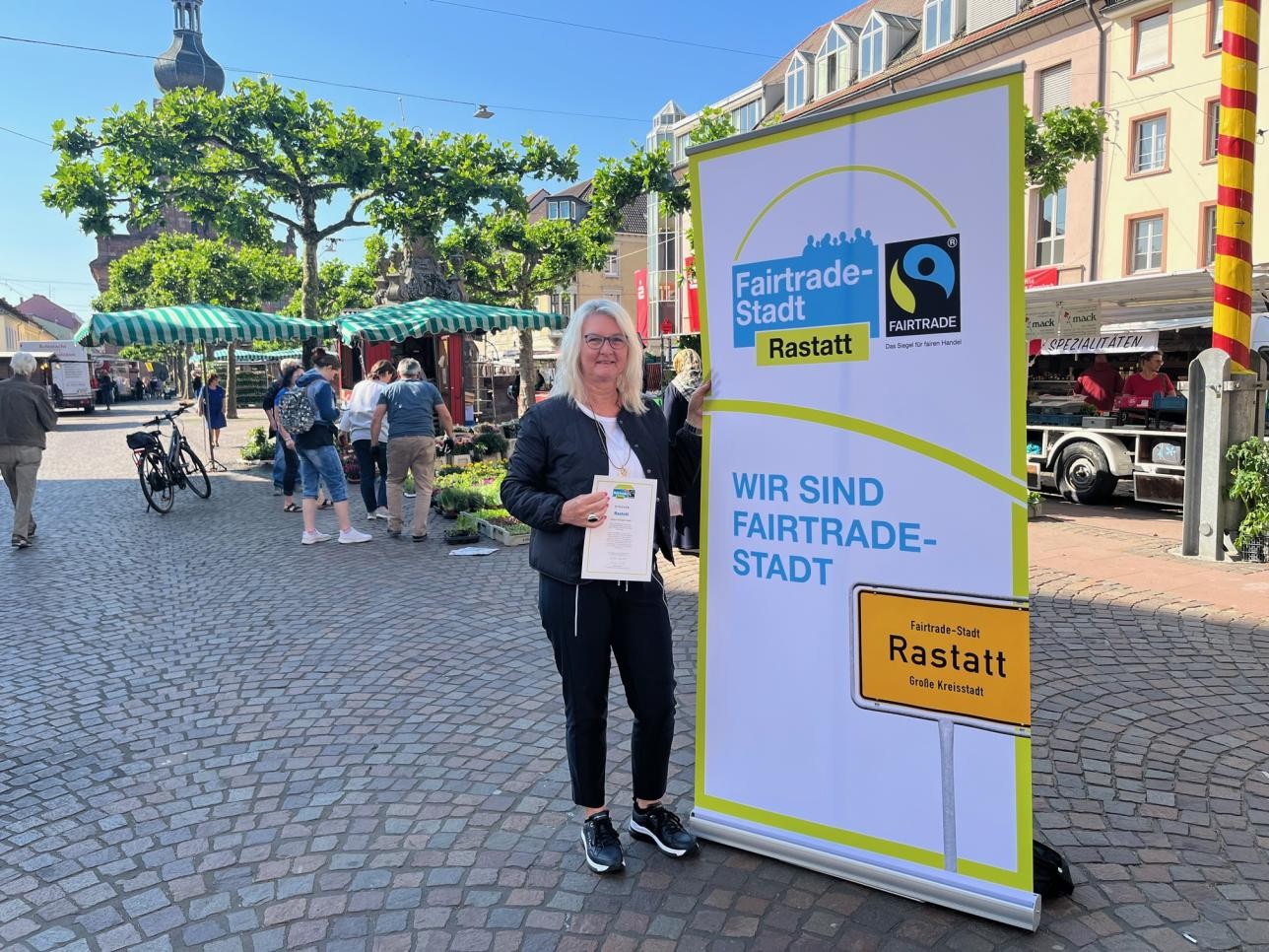 Die neue städtische Koordinatorin, Susanne Jahn, steht auf dem Wochenmarkt und zeigt freudig die Fairtrade-Urkunde. Foto: Heike Bornhäußer/Stadt Rastatt